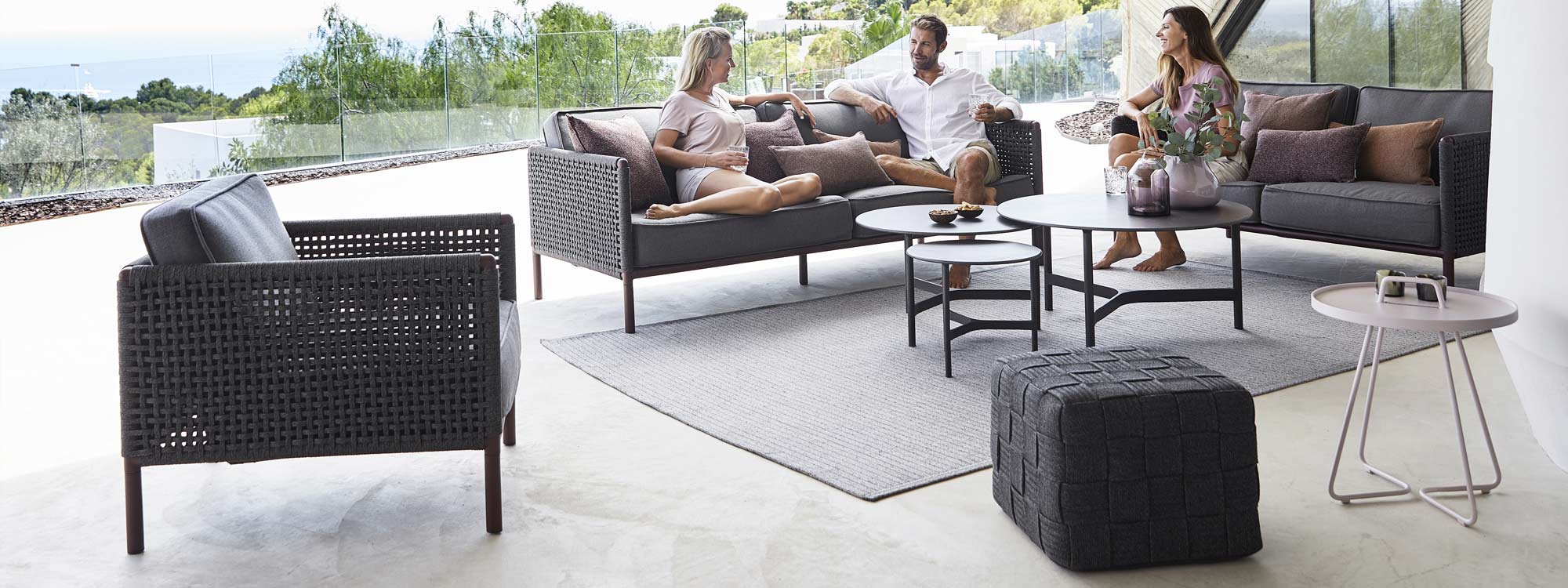 Image of 2 women and a man relaxing on dark grey Encore garden sofa with Cane-line Twist garden low tables in center, shown on minimalist terrace with trees and sea in the background