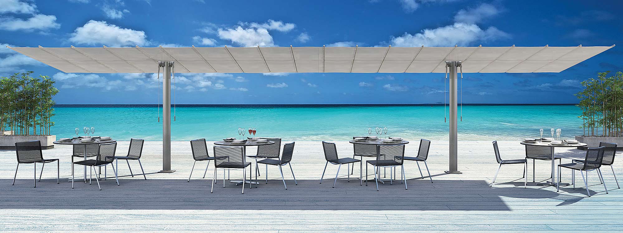 Image of a pair of FIM Flexy Twin free-standing retractable shades placed together, with Coro outdoor furniture beneath, shown on wooden decking with beach and blue sea and sky in the background
