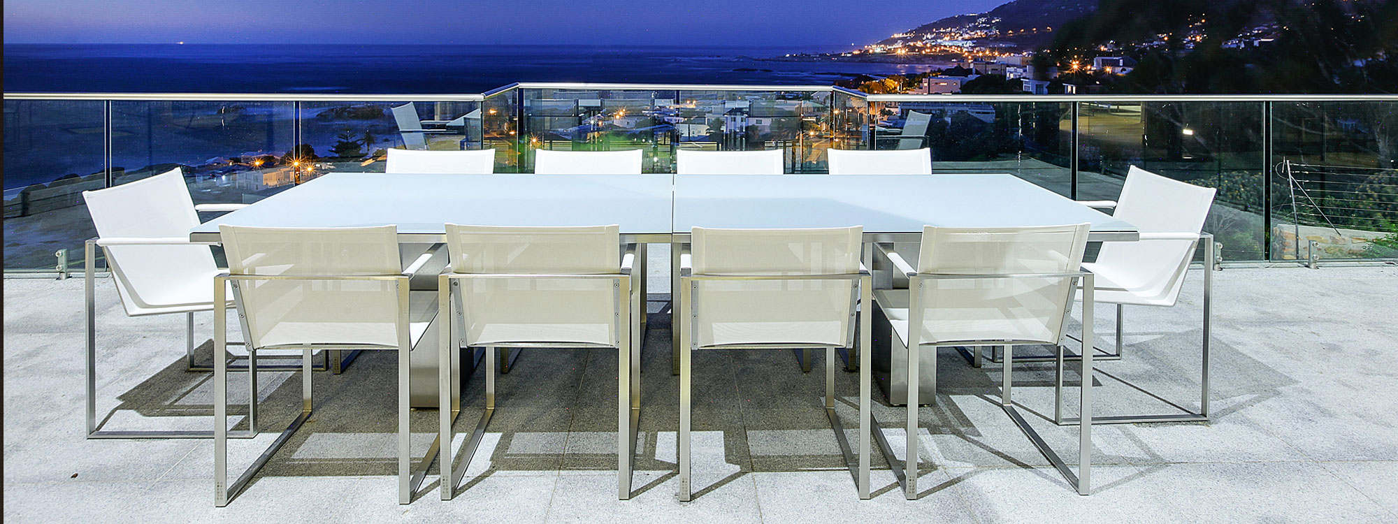 Image of Doble white and stainless steel garden table and Butaque garden chairs, shown on terrace at dusk, with coastline in the background.