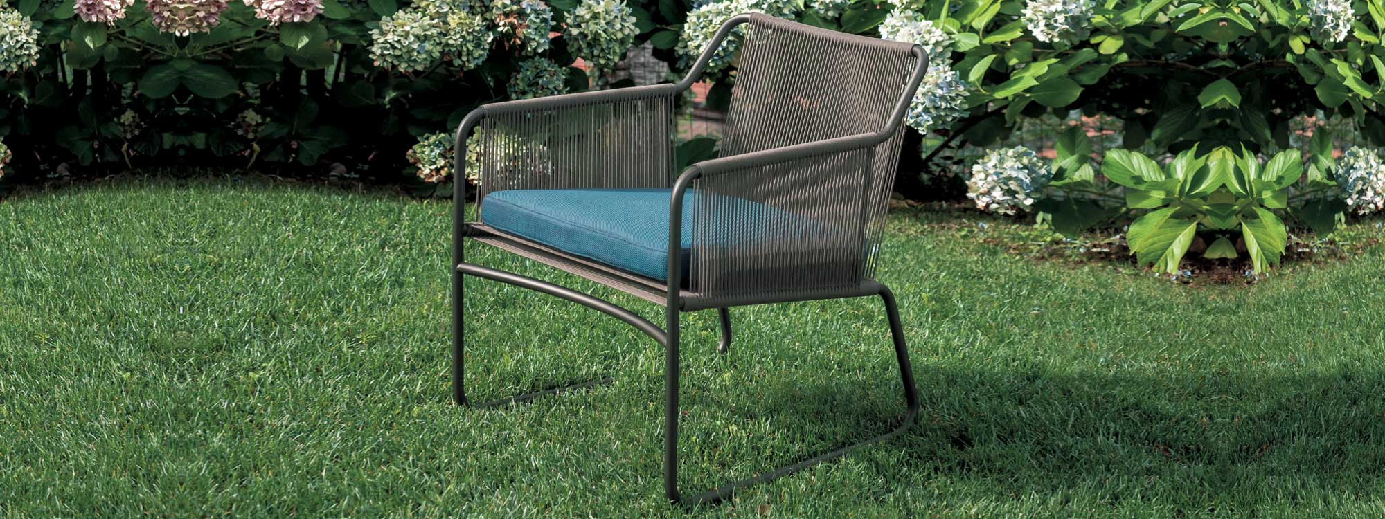 Image of RODA Harp black minimalist garden lounge chair with teal-color cushion, shown on lawn with Hydrangeas in background