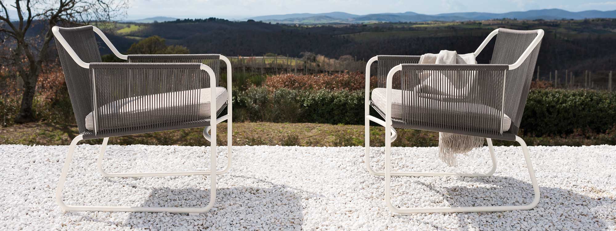 Image of pair of white Harp modern garden lounge chairs with stone colored seat, back and arms on white gravel with Tuscan landscape behind