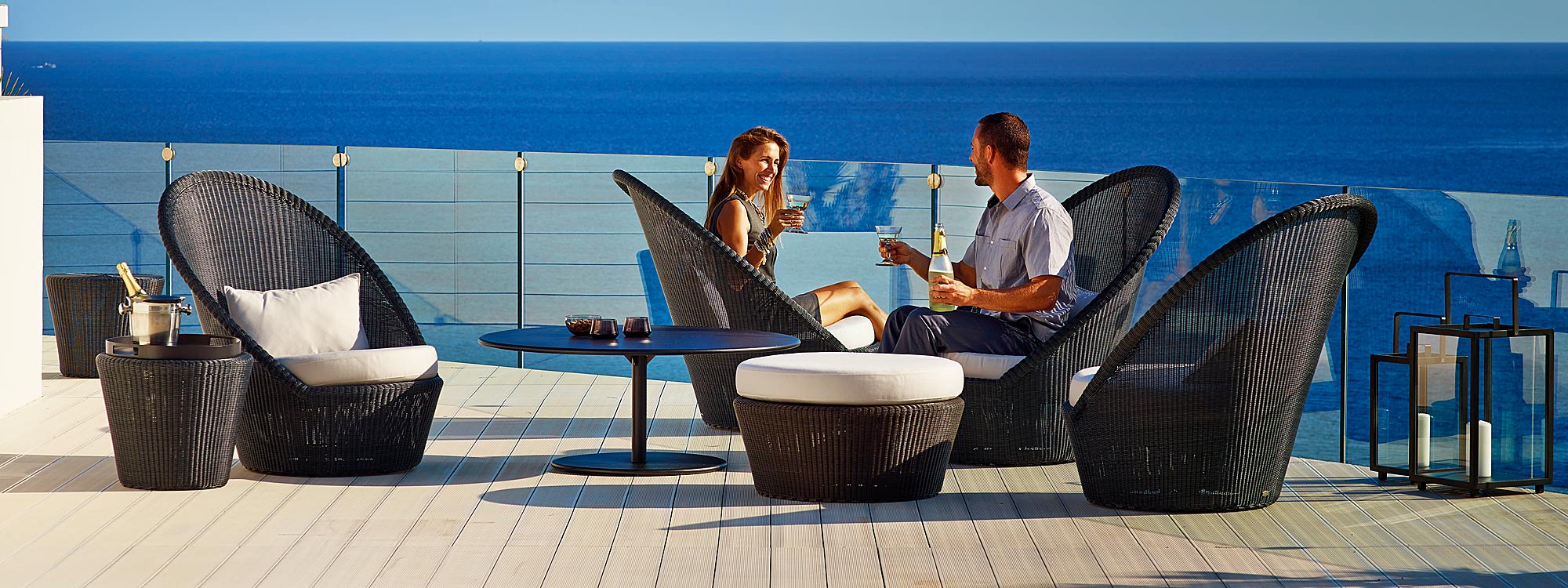 Image of couple enjoying bottle of sparkling wine whilst sat on mocca-coloured Kingston revolving lounge chairs by Caneline garden furniture
