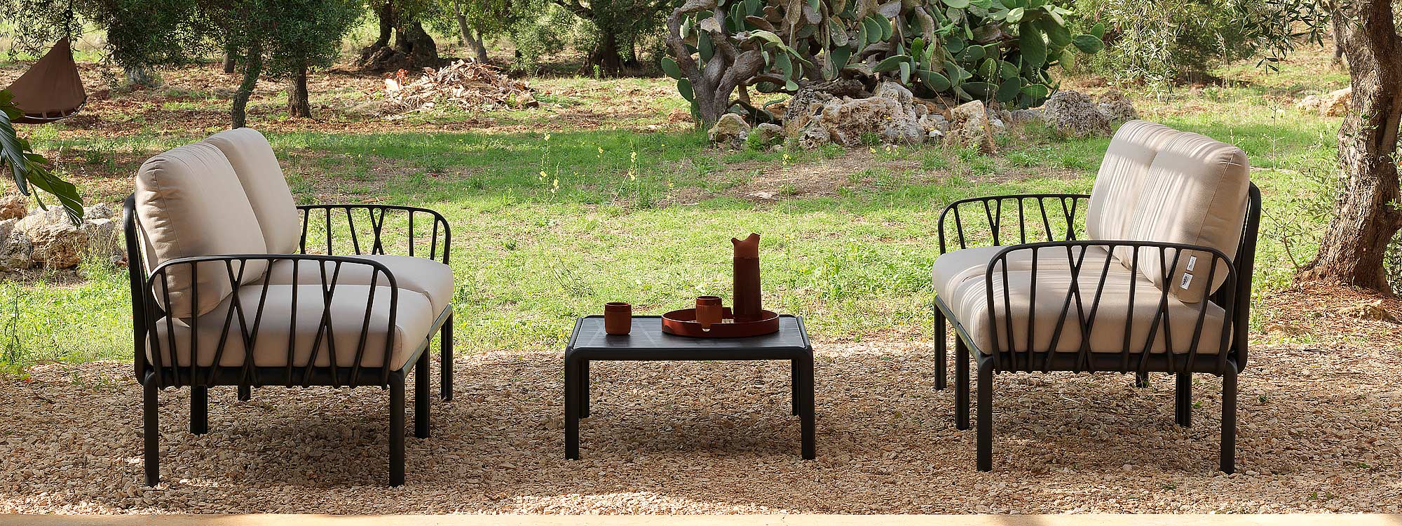 Image of pair of Komodo 2 seat black garden sofas with ecru cushions and low table, shown on wood chip floor with arid and rocky and in the background interspersed with cactuses