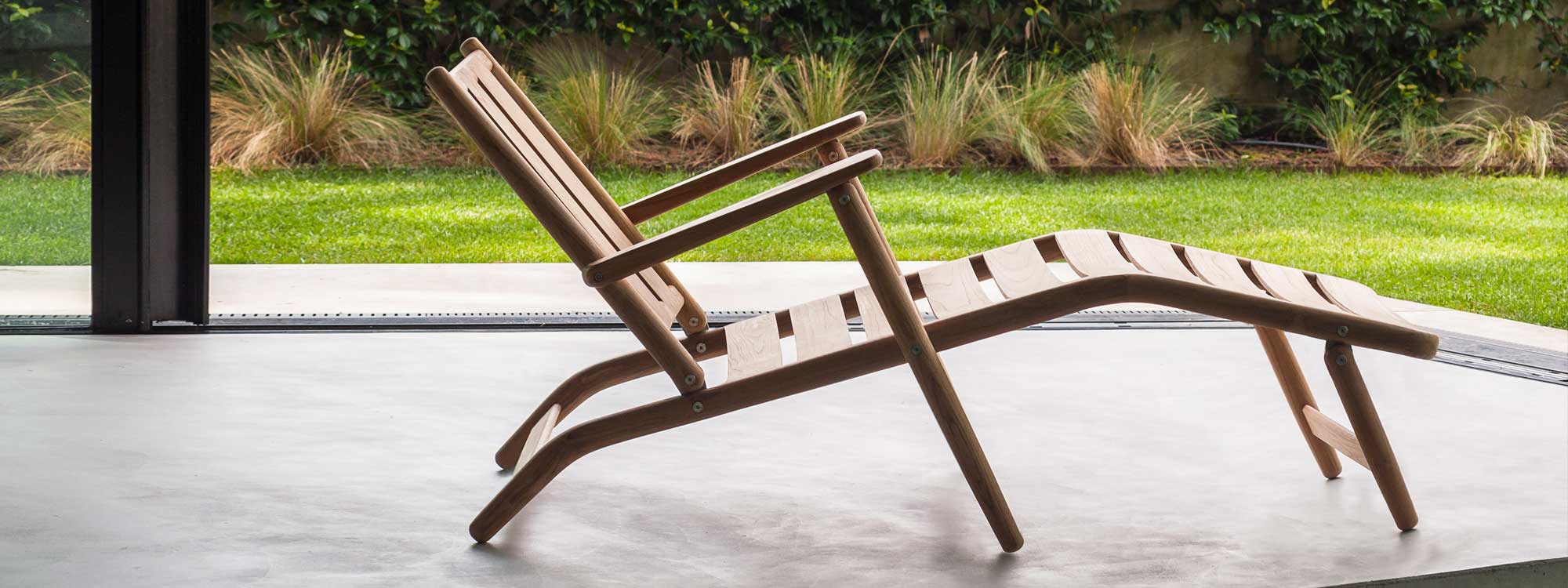 Image of side view of RODA Levante teak steamer chair on poured concrete floor with lawn and architectural grasses in background