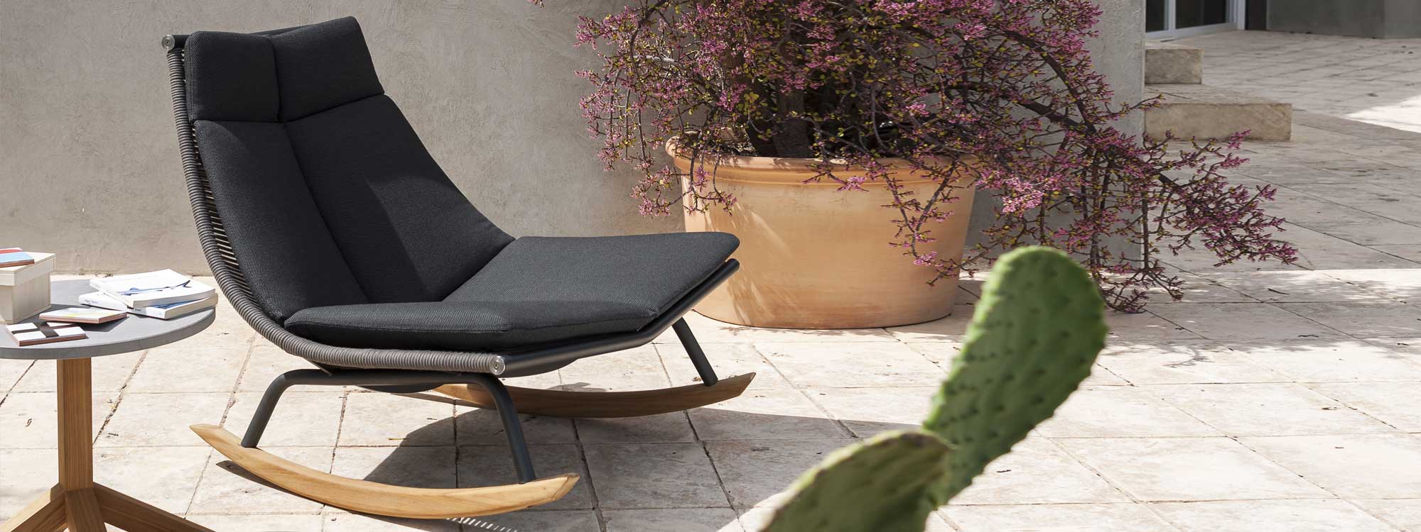 Image of RODA Laze modern outdoor rocking chair with teak skids and black cushion next to Root teak side table, with cactus in foreground
