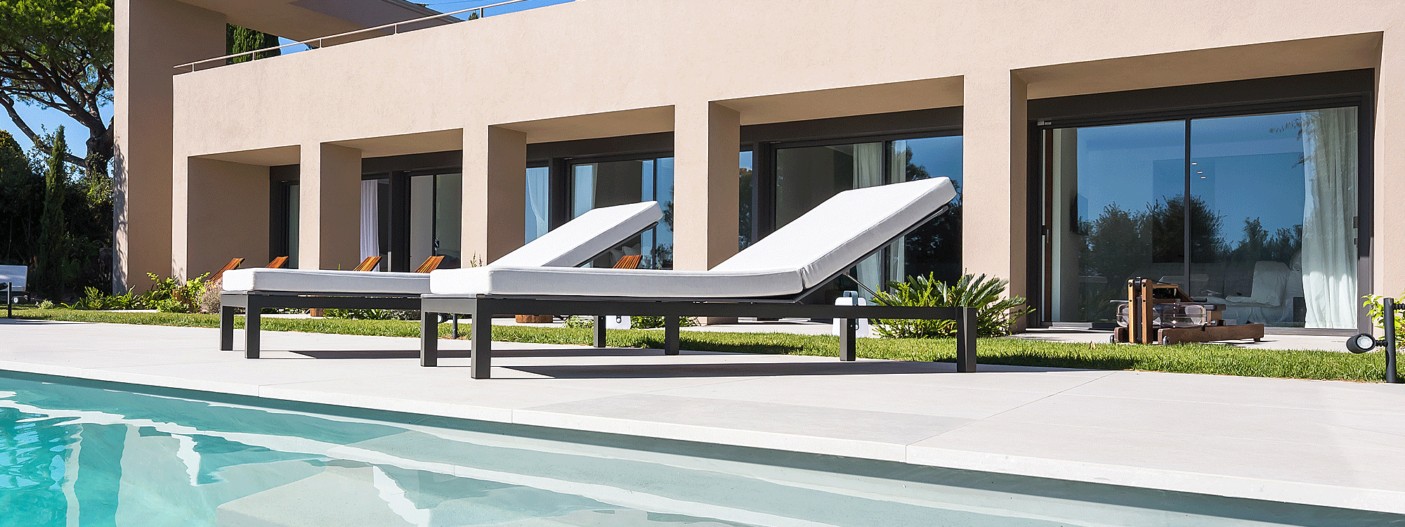 Image from side of 2 Leuven anthracite sunbeds with white cushions on poolside, with hotel rooms and lawn in background