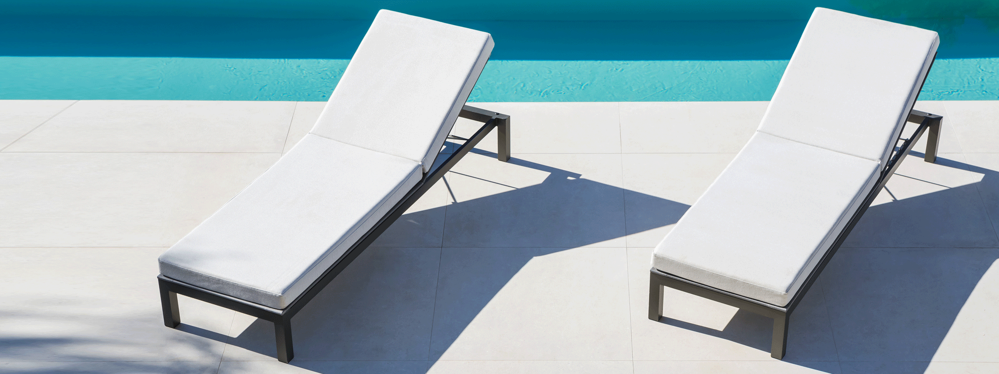 Image of pair of Leuven adjustable sun beds with anthracite frames and white cushions next to still waters of swimming pool