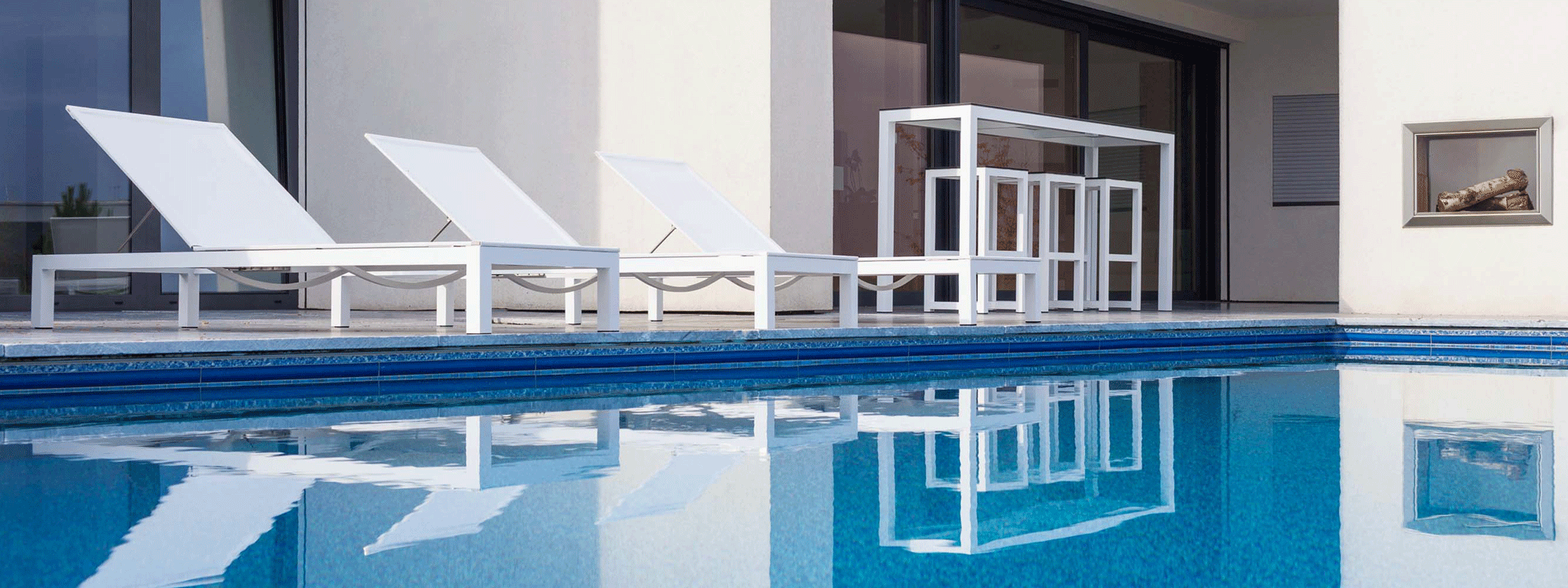 Image of 3 Leuven white sun loungers and white bar table and bar stools on poolside with reflection of furniture shown in the water