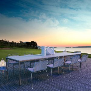 Image of Coro Jubeae stainless steel garden chairs with white woven seat and back, shown around long garden dining table on a terrace at dusk