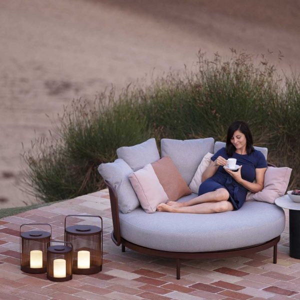 Woman relaxing on Baza daybed next to 3 Rust-Brown coloured Luci LED garden lanterns