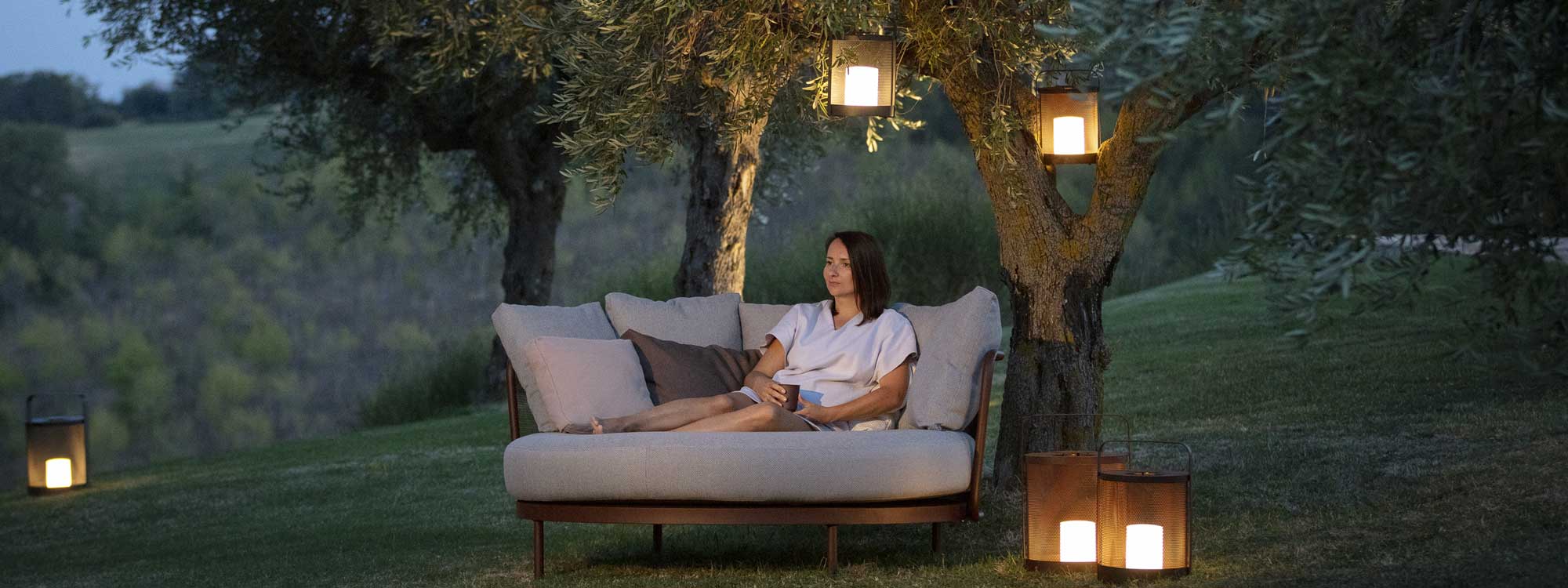 Image of woman sat holding drink in Baza daybed at dusk, beneath tree with lit Luci garden lanterns hanging from branches