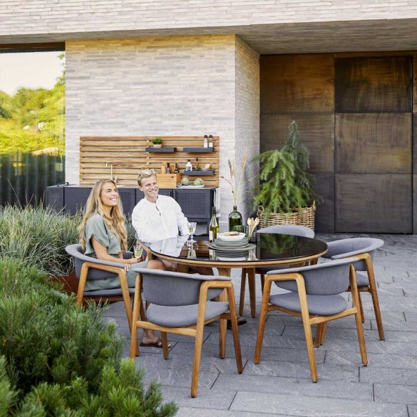 Image of couple sat in Caneline Luna upholstered teak chairs, placed around Aspect round teak table with Smokey Black glass top