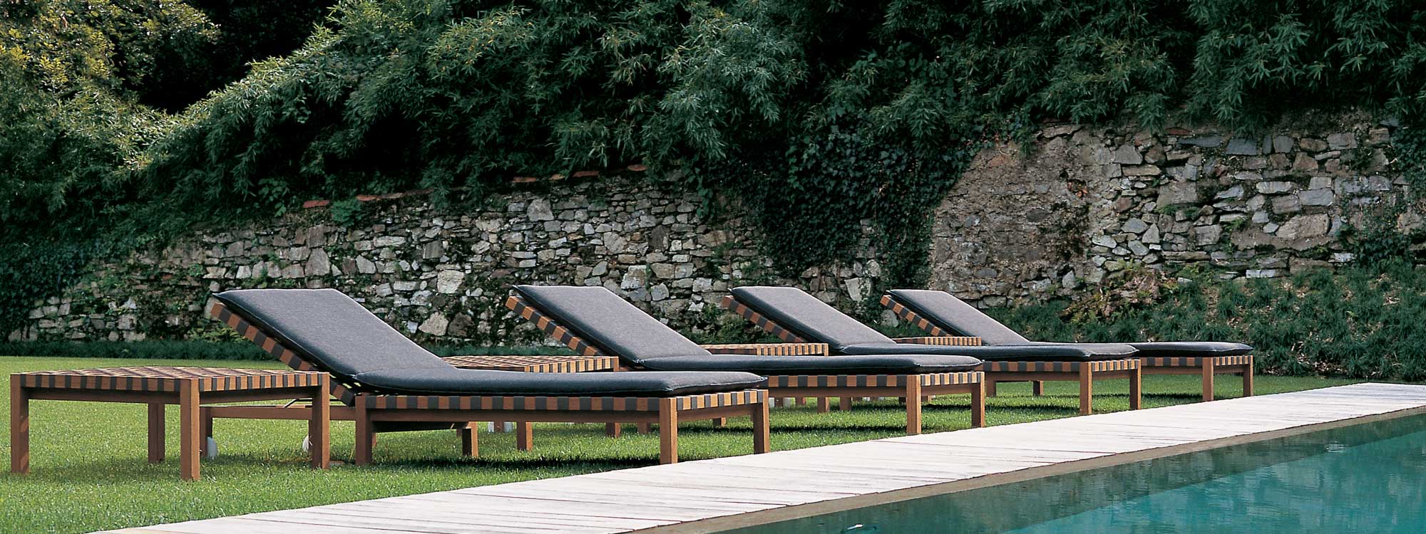 Image of row of 4 RODA Mistral teak sun beds with grey webbing and grey cushions on sunny poolside with lawn and trees in background