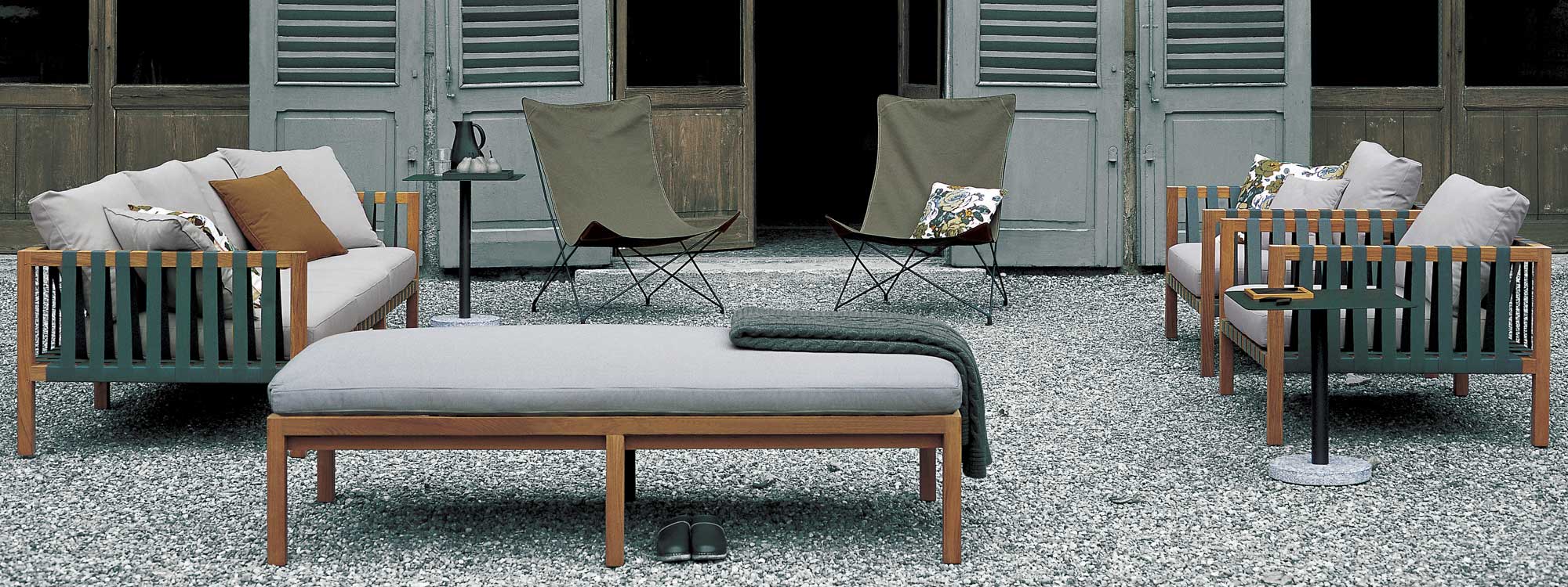 Image of RODA Mistral teak sofa and lounge chairs with teal webbing and light-grey cushions, next to Lawrence butterfly chairs, shown on gravel terrace