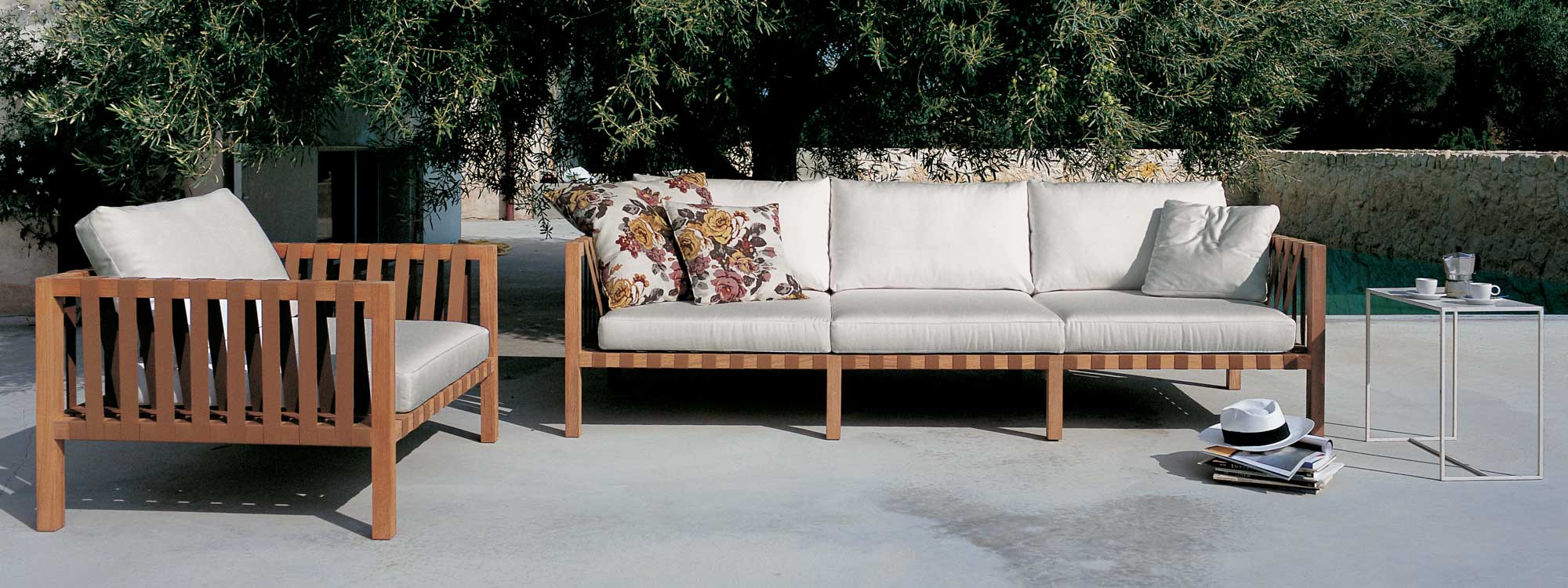 Image of RODA Mistral teak sofa and lounge chair with tobacco-colored webbing and white cushions on sunny terrace with tree in background