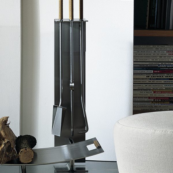Image of Peter Maly stainless steel fireside accessories with stand by Conmoto, on hearth next to stacked books