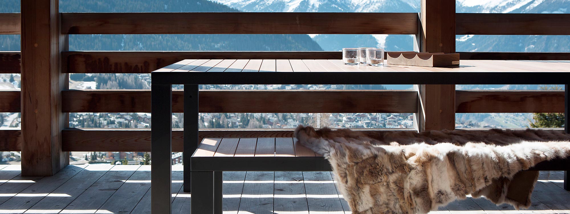 Image of Roshults garden dinner table and bench seat with fur throw, shown on chilly balcony with snow-capped mountains in the background