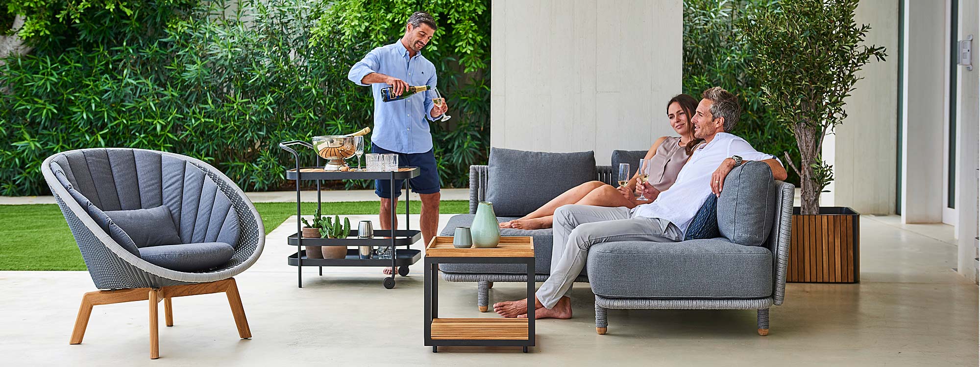 Image of man pouring drinks for guests sat on Moments grey corner sofa and Peacock lounge chair by Cane-line