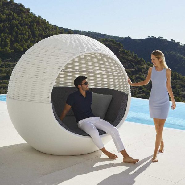 Image of man sat inside Moon round daybed on sunny terrace whilst chatting to woman stood to the side
