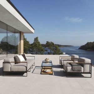 Image of Moore minimalist outdoor lounge furniture on sunny terrace, with modern building with floor to ceiling windows to the left and woodland and lake in the background
