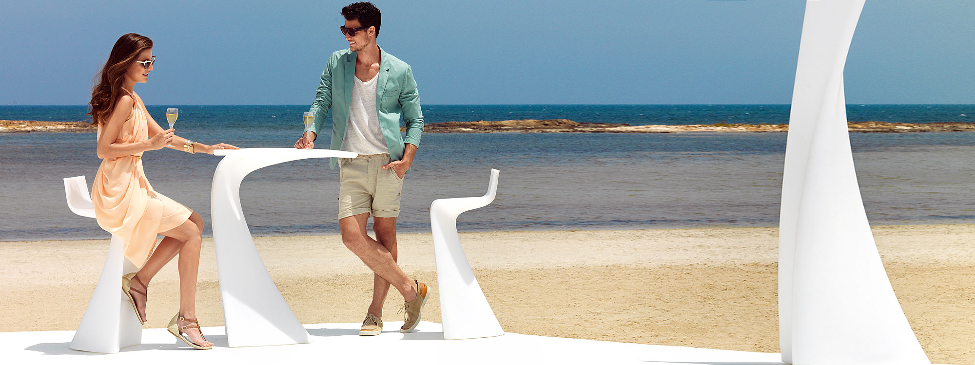 Image of woman and man sat and stood at Vondom Wing white bar table and bar stools on sunny beach front with blue sea and sky in the background