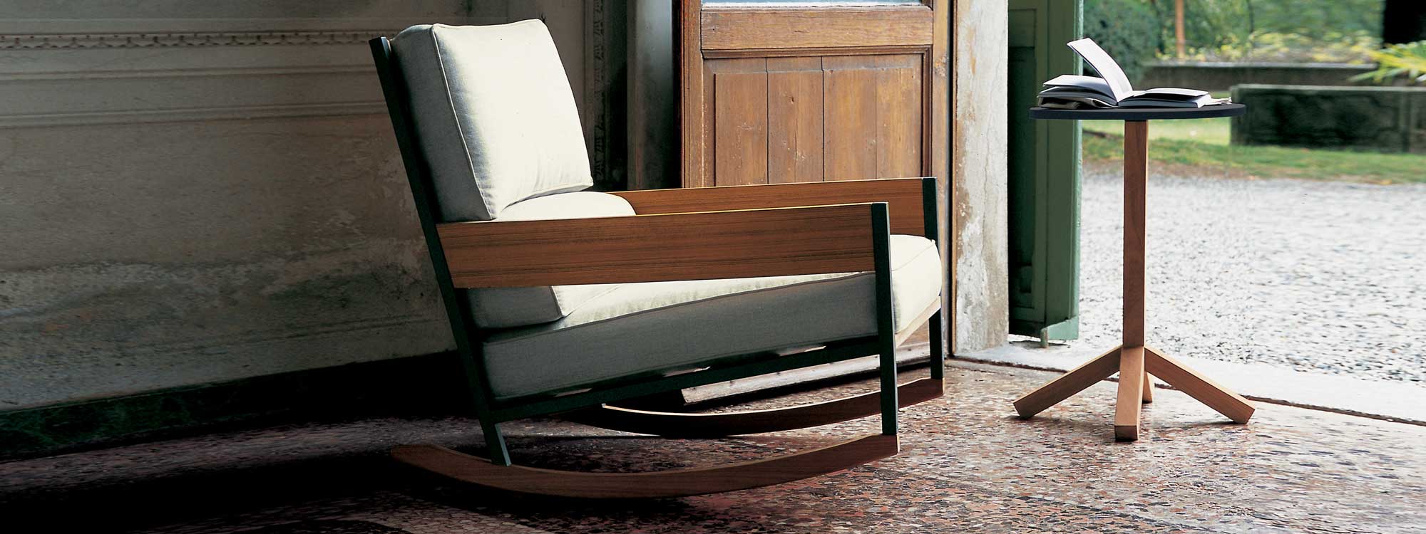 Interior image of RODA Nap rocking chair in teak and smoke-colored stainless steel and white cushions, next to Root side table
