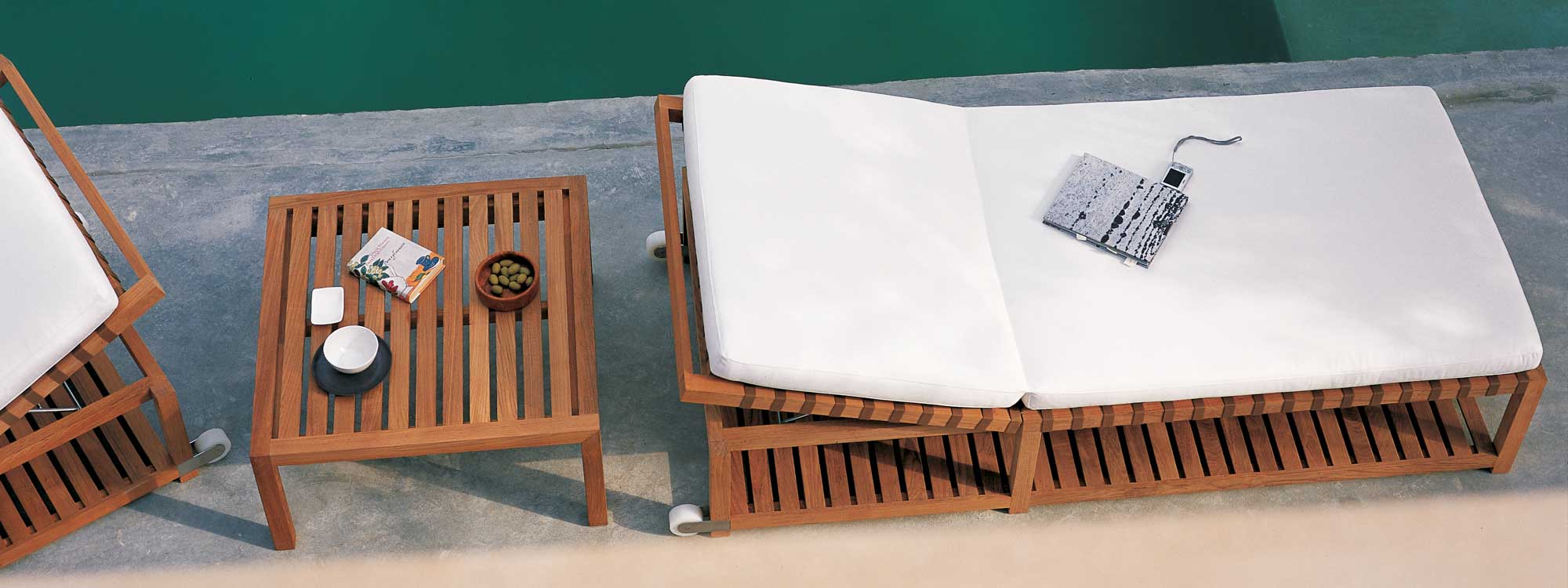 Image of pair of RODA Network minimalist teak loungers on poolside, with teak low table in between with bowl of olives and book on top