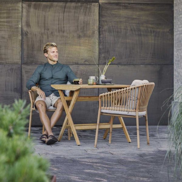 Image of man sat in Caneline Ocean bamboo garden chair next to Flip folding teak table
