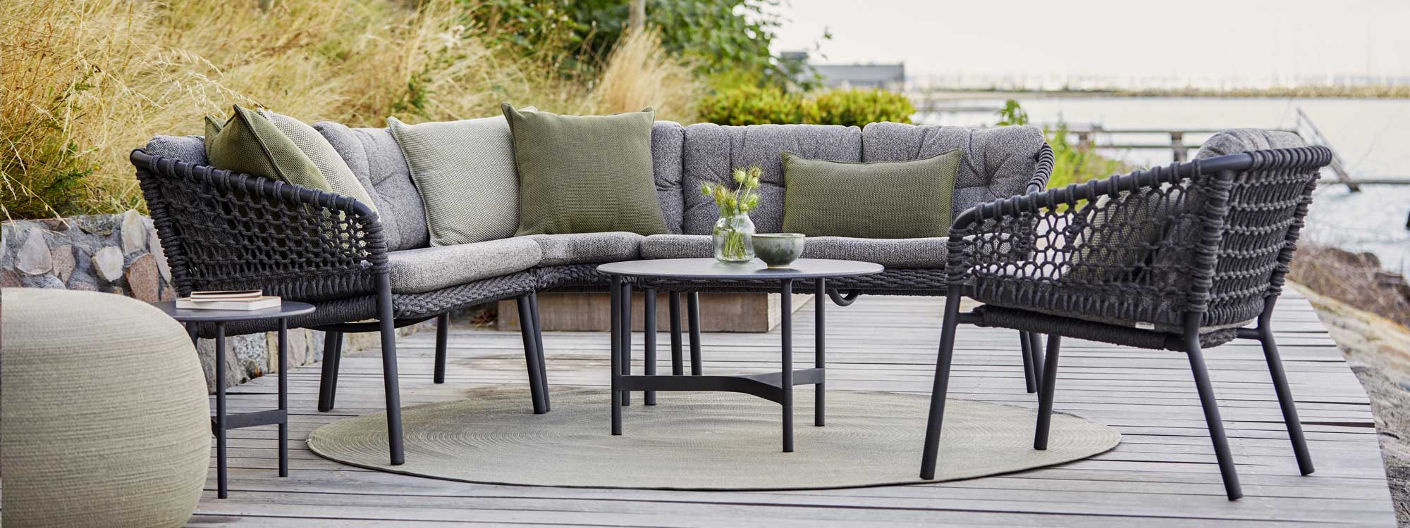 Image of grey Ocean sofa and grey Twist coffee table by Caneline, shown on decking beside grasses and the sea