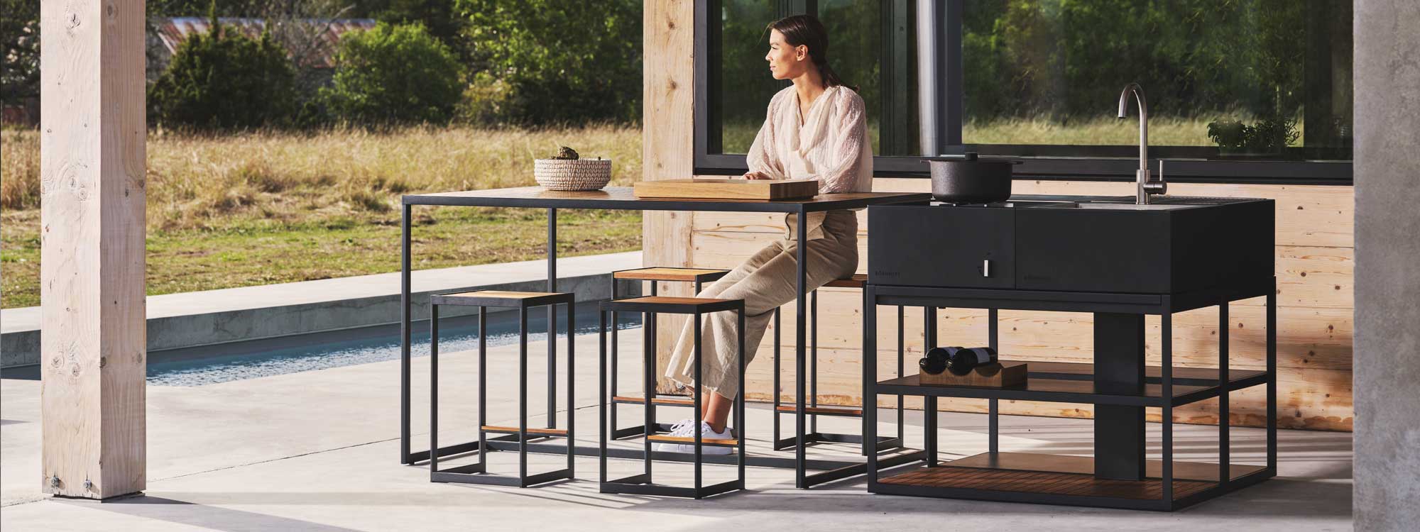 Image of woman sat at Open Bistro outdoor kitchen furniture next to Open Kitchen outdoor bbq and sink by Roshults, shown on minimalist garden terrace