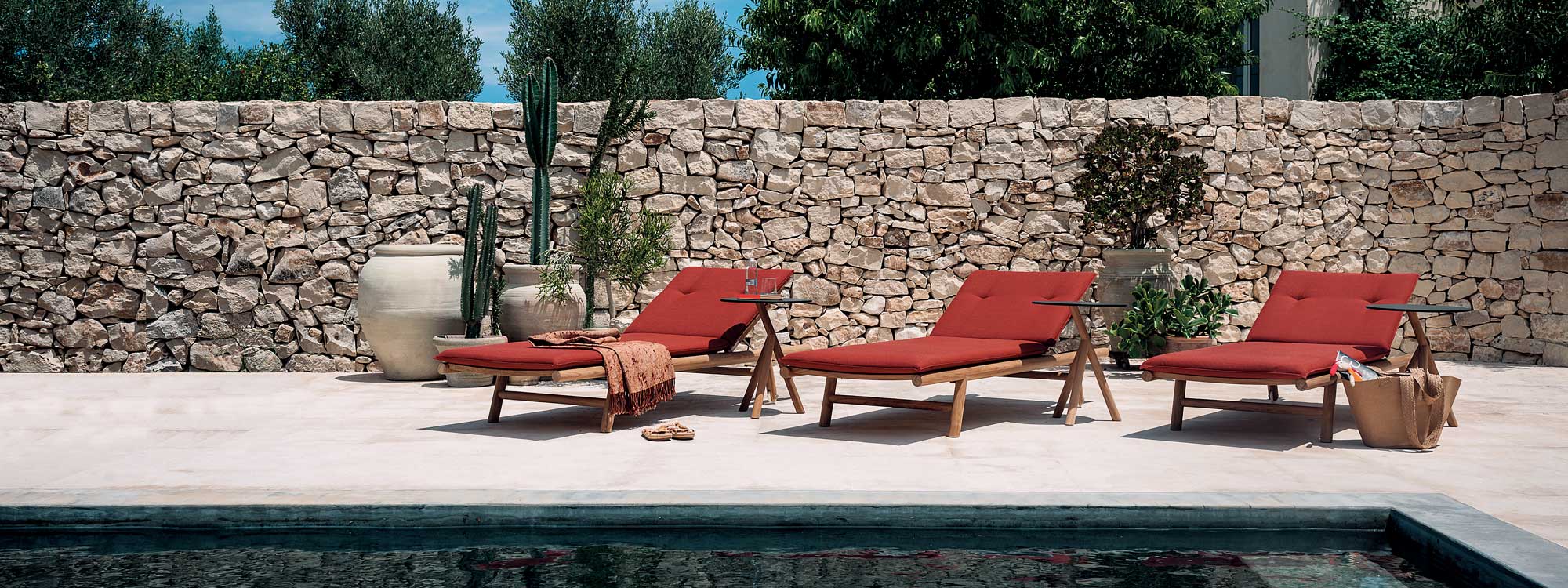 Image of row of 3 RODA Orson teak sun loungers with read cushions on poolside, with dry stone wall in background