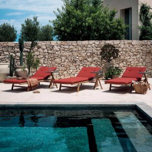 Image of row of 3 Orson modern teak sunbed with red cushions by RODA, shown on sunny poolside with drystone wall in background with tree tops behind
