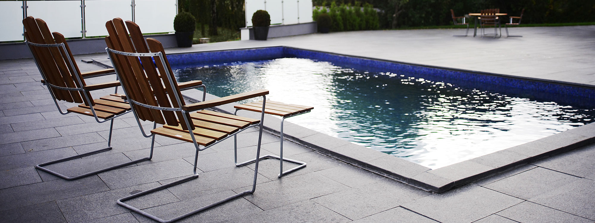 Image of pair of Grythyttan A3 cantilever garden lounge chairs and foots tools with galvanised steel frames and teak surfaces, shown on poolside at dusk
