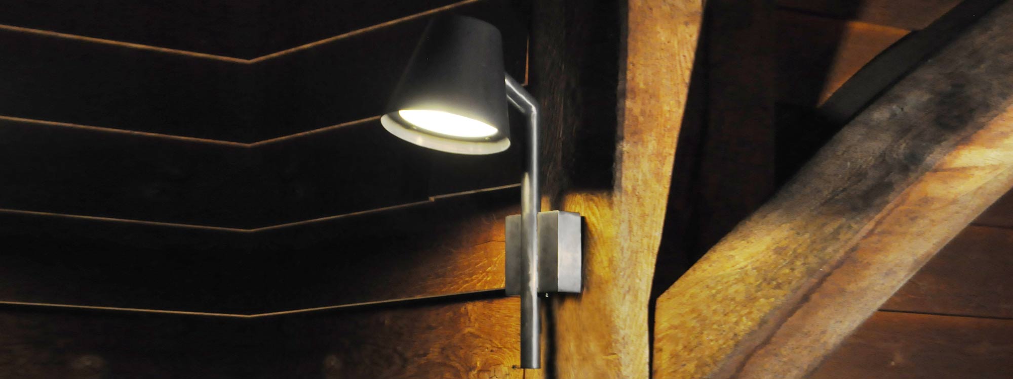 Nighttime image of illuminated Royal Botania Parker wall light in antique brass finish, mounted on wooden weather board