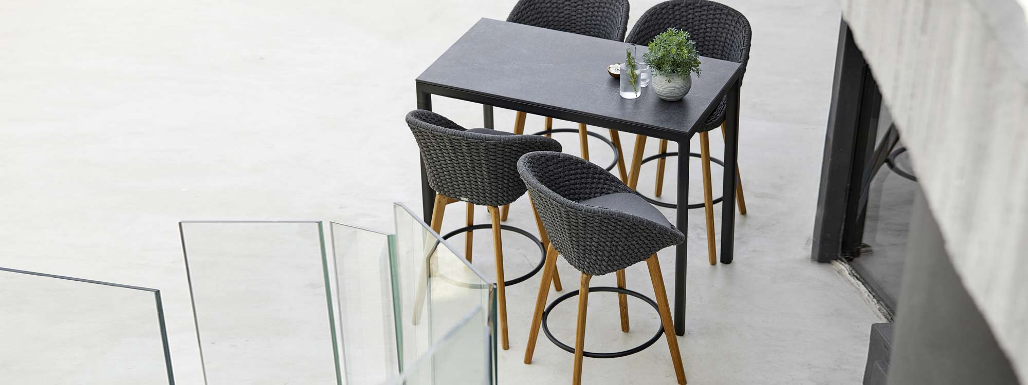 Image of 4 Cane-line Peacock bar stools in dark grey SoftRope with teak legs, shown around Drop high bar table with black Fossil ceramic table top