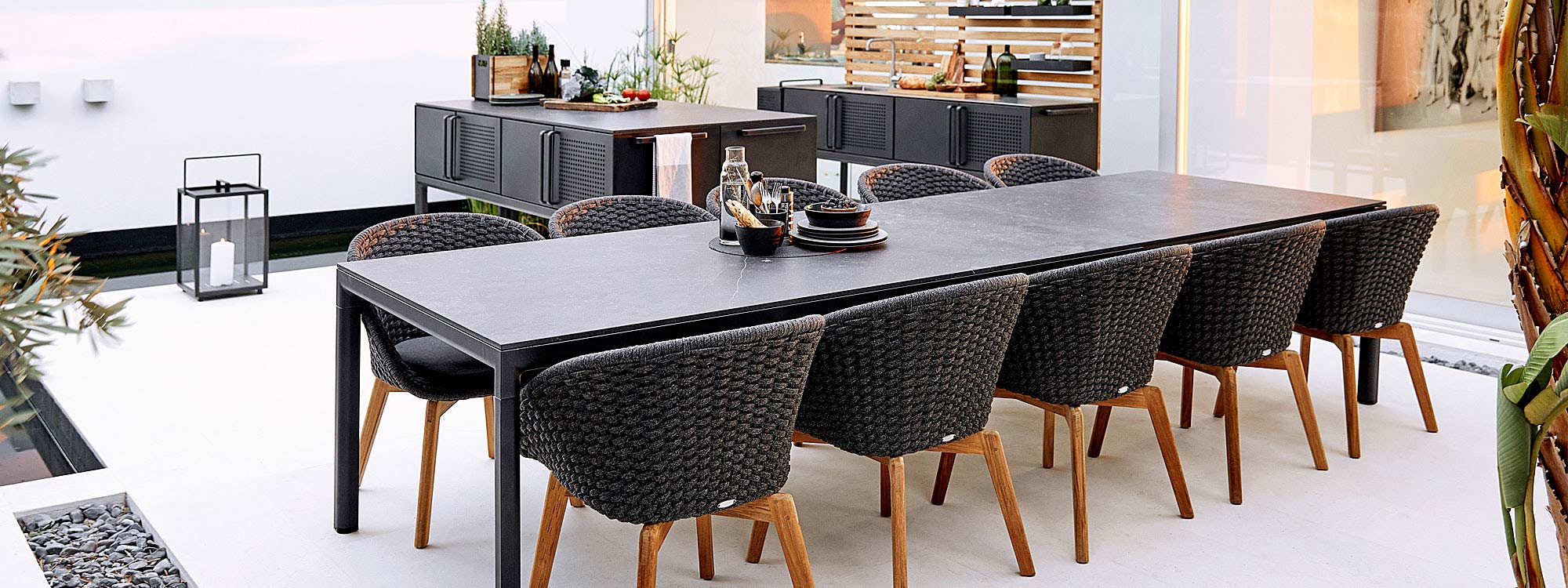 Image of dark-grey and teak Peacock garden chairs and lava-grey Drop ceramic garden table by Cane-line, with Drop outdoor kitchen island in background