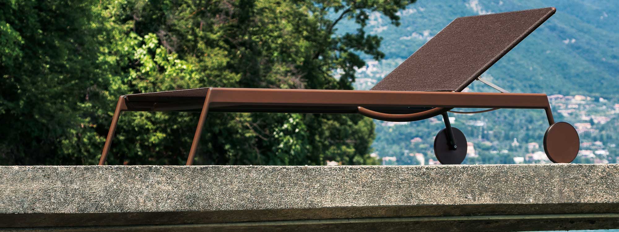 Image of rust-colored Piper modern sun lounger by RODA, with Italian summer hillside in background