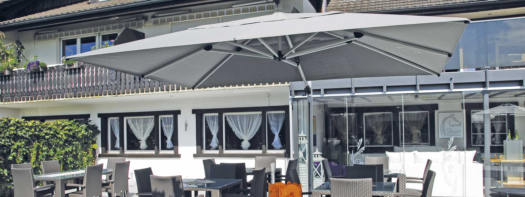 Image of Shademaker Polaris large cantilever parasol with taupe canopy, shown on hotel terrace above bistro furniture