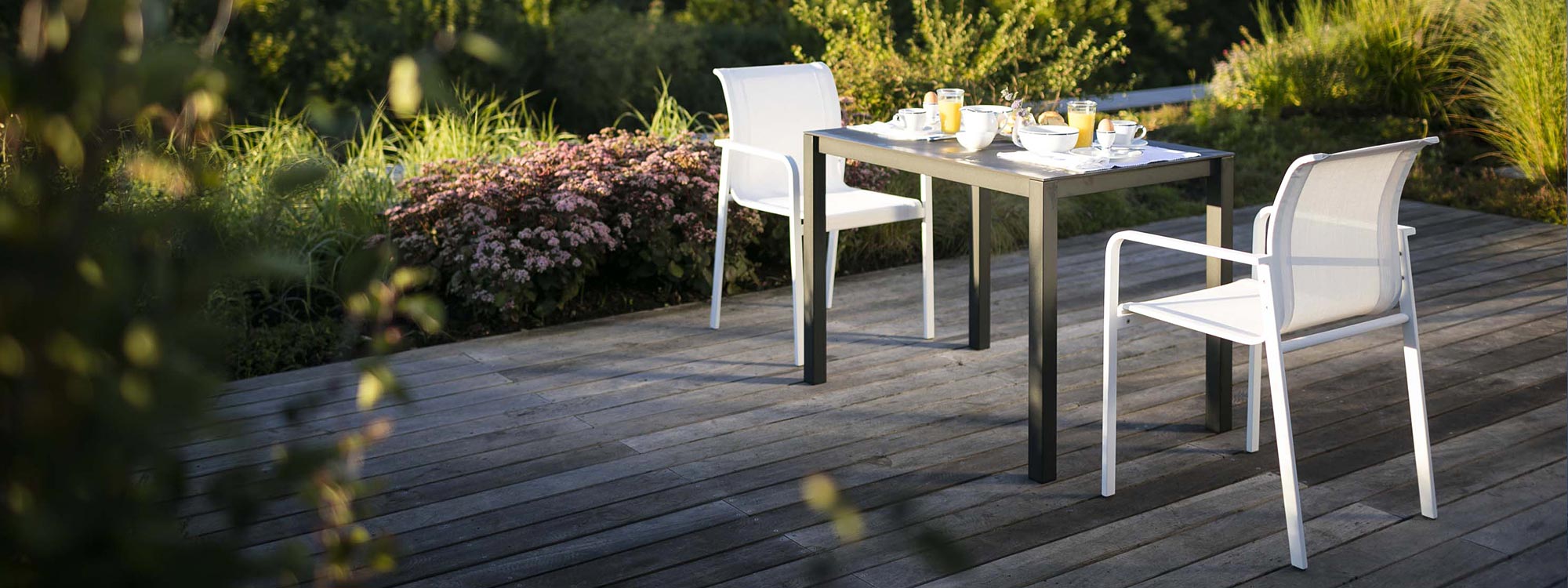 Image of Puro small outdoor dining table and Puro modern garden chair on decking in front of flower bed