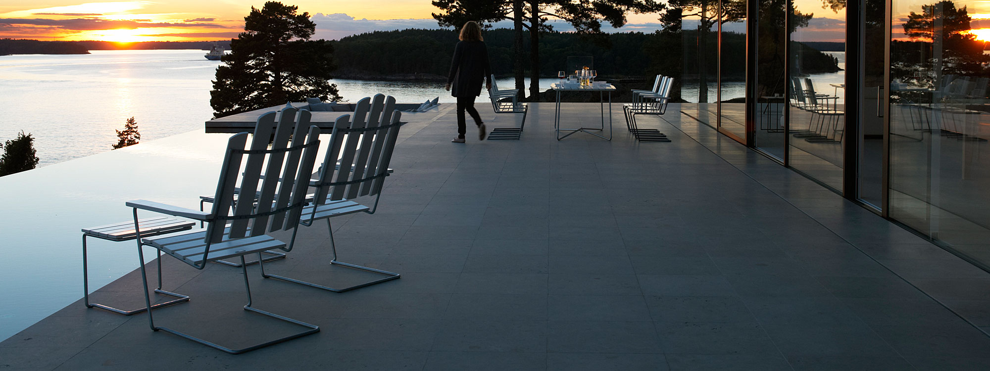 Image of pair of Grythyttan A3 classic garden lounge chairs and foot stools on balcony at dusk, overlooking horizon swimming pool, a lake and woodland