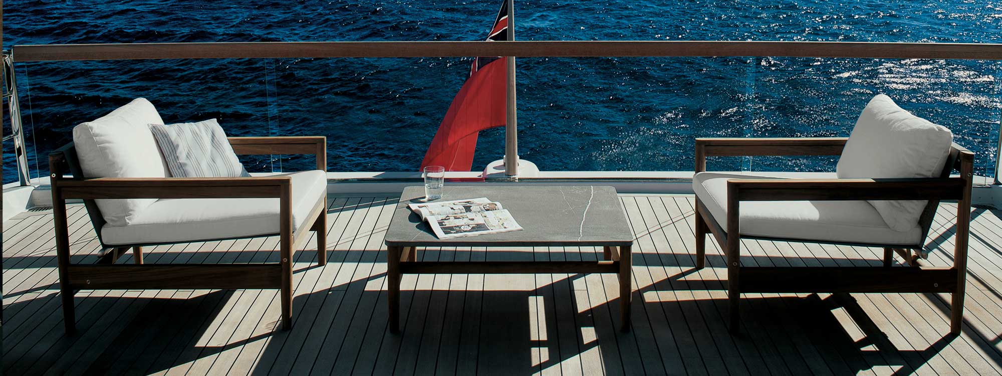 Image of teak aft deck of yacht with RODA Road teak lounge chairs and low table, with fluttering ensign flag and sea in the background