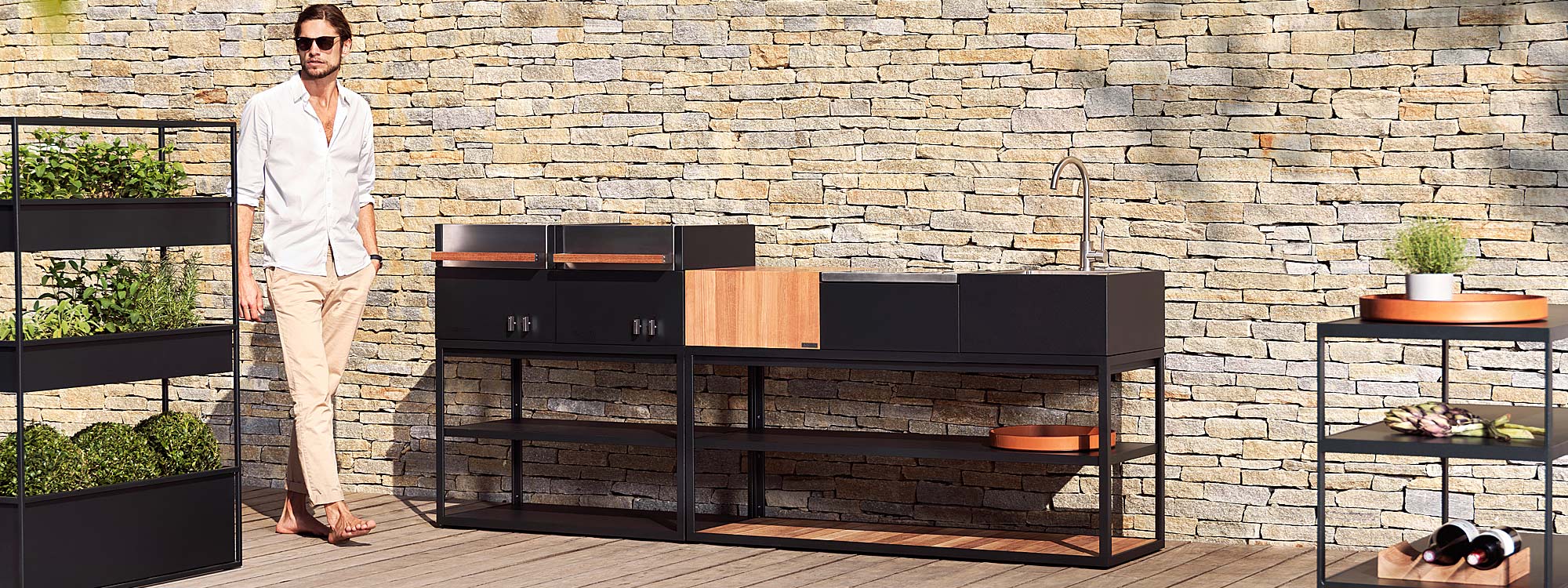 Image of dude in sunglasses stood next to Roshults Open Kitchen modern outdoor kitchen island in anthracite coloured stainless steel and teak, shown in sunny courtyard