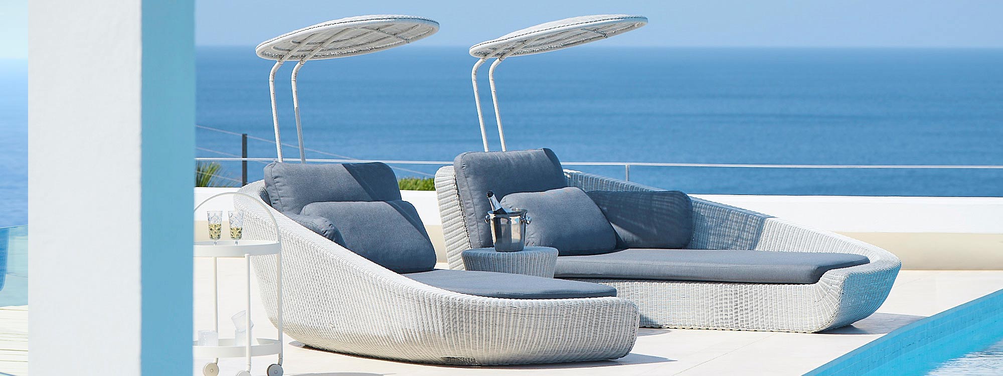 Image of pair of White-grey Savannah woven outdoor daybeds with grey cushions by Cane-line, shown on sizzling terrace with sparkling sea in background