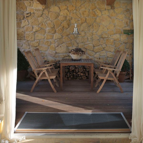 Image of RiZZ The New Standard silver aluminium & teak doormat in doorway, with teak dining furniture and stone wall in the background