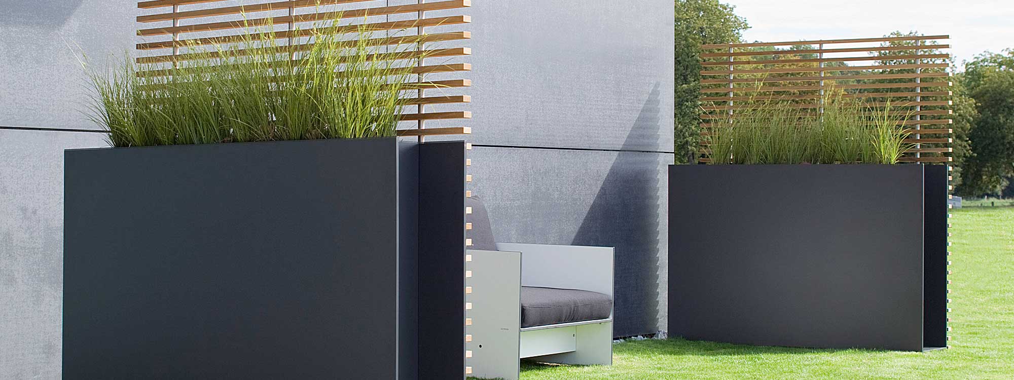 Image of pair of Sotomon anthracite raised planters with ash trellis on a lawn against a poured concrete wall, with a white Riva garden lounger in between the planters