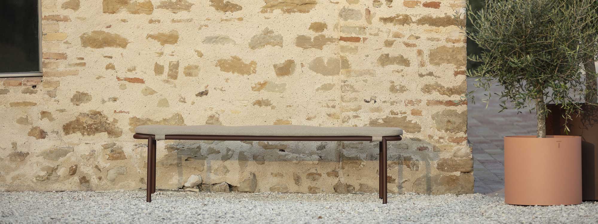 Image of rust-brown colored Starling garden bench with taupe seat cushion, shown on gravel against stone wall