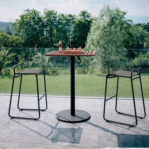 Image of RODA Stem tall bistro table with Harp modern garden bar chairs, shown on terrace with lawn and shrubs in background