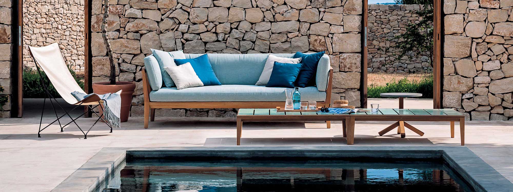 Image of RODA Teka 3 seat teak sofa with light blue cushions, together with Teka teak low table with glazed ceramic top, shown next to tranquil water feature with drystone wall in background