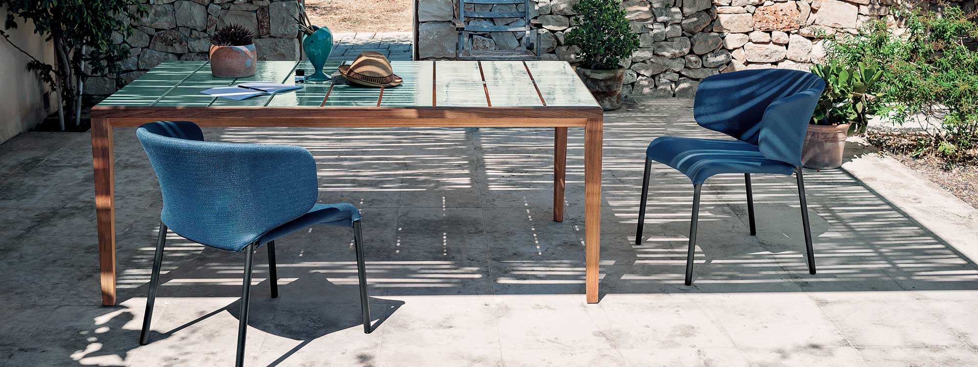 Image of RODA Teka teak dining table with top in slats of glazed ceramic, next to Double upholstered garden chairs in blue fabric.