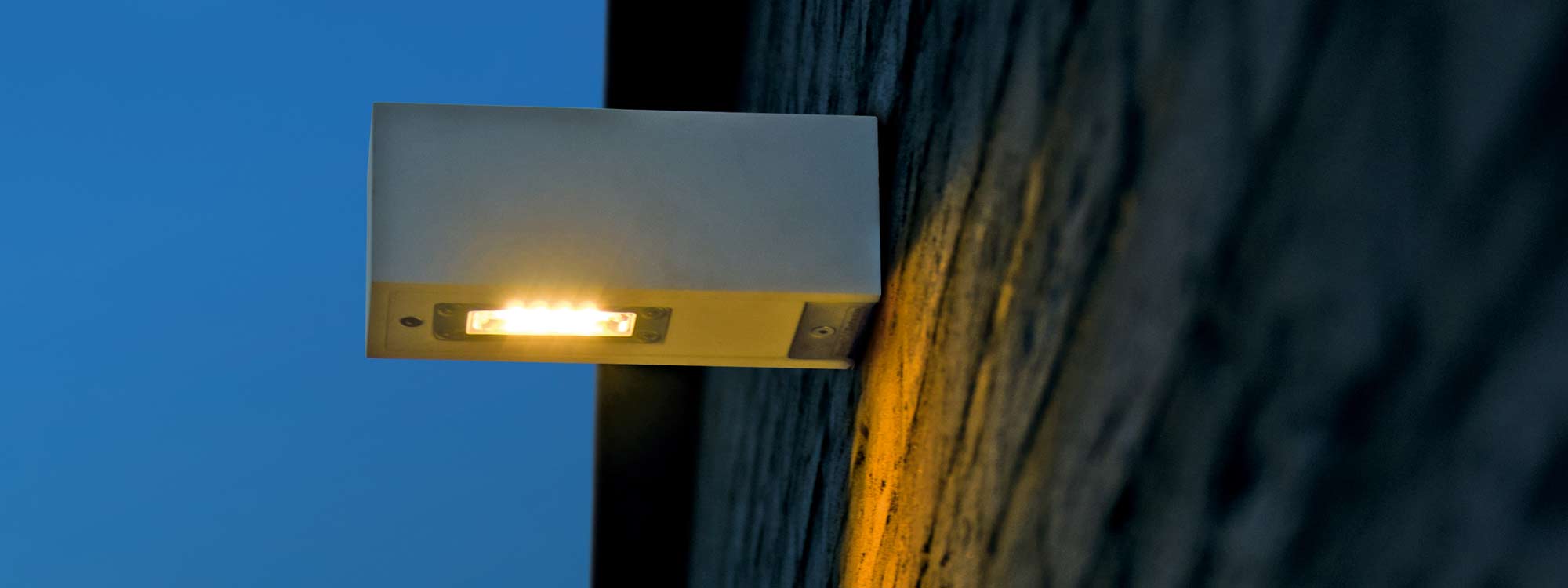 Image of Royal Botania Twin Wall up or down light in white corian which diffuses a warm glow, shown on brick wall at dusk